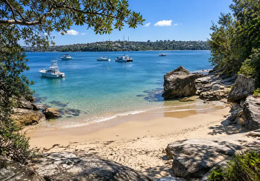 Flat, clear harbour water and a small strip of sand below sandstone boulders and bushland at Castle Rock Beach, Clontarf